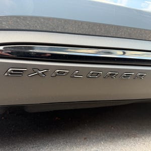 May include: A matte grey automotive exterior panel featuring the word EXPLORER in raised, metallic silver lettering. Above the text, a horizontal, glossy black trim piece runs across the surface, reflecting light along its curved edge.