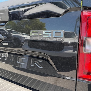 May include: Close-up of a black Ford Maverick truck bed. The Ford logo is visible, along with the text "F-150 LIGHT DUTY" in silver lettering. The Maverick name is also displayed in silver.