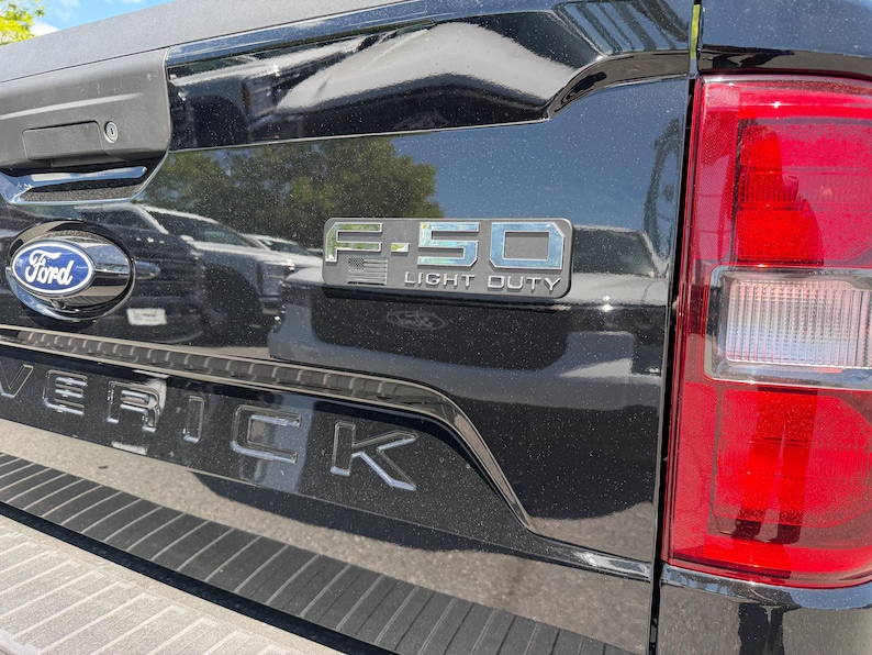 May include: Close-up of a black Ford Maverick truck bed. The Ford logo is visible, along with the text "F-150 LIGHT DUTY" in silver lettering. The Maverick name is also displayed in silver.