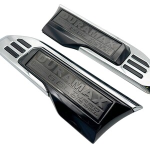 May include: Two black and chrome Duramax 6.6L Turbo Diesel truck accessories. The accessories have the text "DURAMAX 6.6L TURBO DIESEL" embossed on a black surface. The accessories have a chrome border and three black slots.