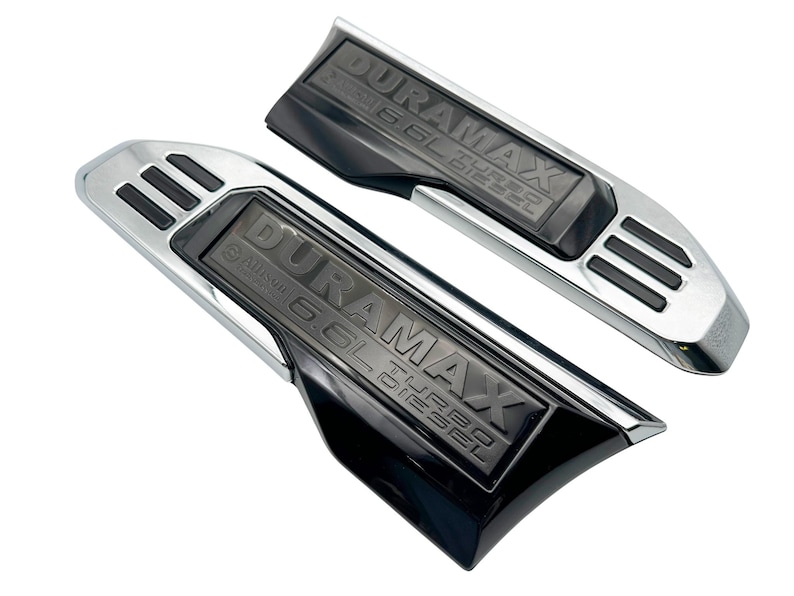 May include: Two black and chrome Duramax 6.6L Turbo Diesel truck accessories. The accessories have the text "DURAMAX 6.6L TURBO DIESEL" embossed on a black surface. The accessories have a chrome border and three black slots.