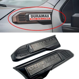 May include: Black and brown Duramax emblem for a vehicle. The emblem has the text "DURAMAX" in white, with "Allison" and "6.6 LITER" in red and white. The emblem is shown installed on a vehicle and also as a pair of emblems.