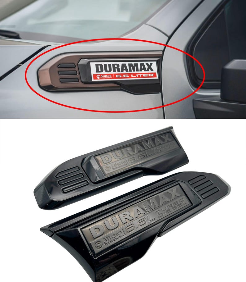 May include: Black and brown Duramax emblem for a vehicle. The emblem has the text "DURAMAX" in white, with "Allison" and "6.6 LITER" in red and white. The emblem is shown installed on a vehicle and also as a pair of emblems.