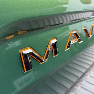 May include: Close-up of a green vehicle's body with the chrome and orange "MAVERICK" emblem. The letters have a black outline and are set against a glossy green surface.