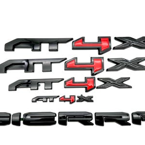 May include: Black and red "AT4X" and "SIERRA" emblems for a truck. The emblems are made of a glossy black plastic with red accents.