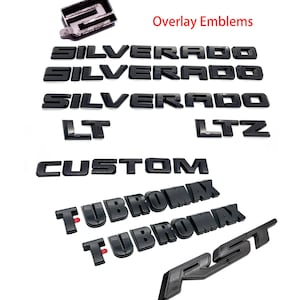 May include: Black overlay emblems for vehicles, including "Silverado," "LT," "LTZ," "Custom," "TubroMax," and "RST." The text "Overlay Emblems" is also visible. The emblems are in a glossy finish.