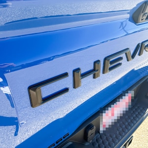 May include: Close-up of a blue vehicle's rear with the black "CHEVROLET" emblem. The emblem is outlined in black. The vehicle's bumper and a portion of the license plate are visible.