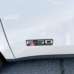 May include: A white vehicle's side panel with a black and silver emblem. The emblem reads "F-150 LIGHT DUTY" and features a small American flag. The tire and part of the vehicle's body are visible.