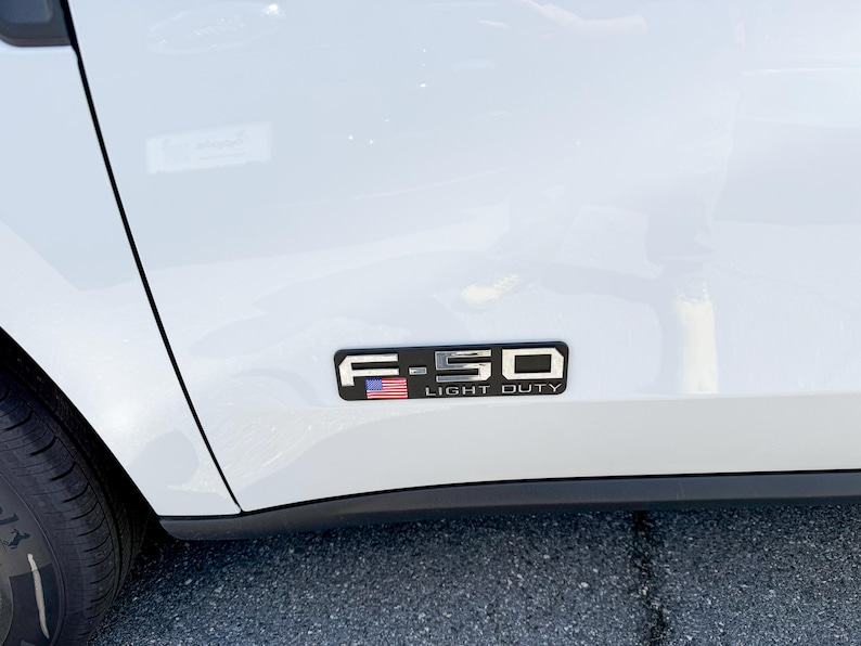 May include: A white vehicle's side panel with a black and silver emblem. The emblem reads "F-150 LIGHT DUTY" and features a small American flag. The tire and part of the vehicle's body are visible.