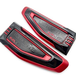 May include: Two automotive emblems featuring a glossy black and matte red finish. Each piece displays the embossed text ULTIMATE and 6.2L V8. The design includes three horizontal slats on one end and a tapered, angular shape with raised borders.