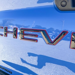 May include: Close-up of a blue vehicle's tailgate with the chrome "CHEV" emblem outlined in red. The word "COLORADO" is visible at the bottom. The image is taken in daylight, with reflections on the shiny surface.