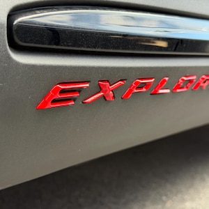 May include: A matte dark grey automotive panel featuring the word EXPLORER in bold, glossy red lettering. Above the text, a smooth, horizontal black trim piece with a reflective surface is visible, creating a contrast against the textured grey finish.