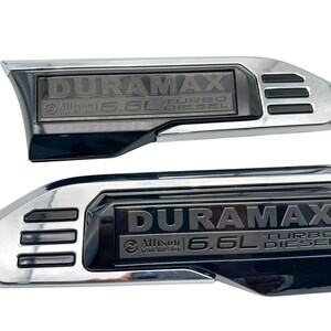May include: Two chrome and black Duramax Turbo Diesel emblems. Each emblem features the text "DURAMAX", "Allison", and "6.6L TURBO DIESEL". The emblems have a curved shape with a rectangular centre.