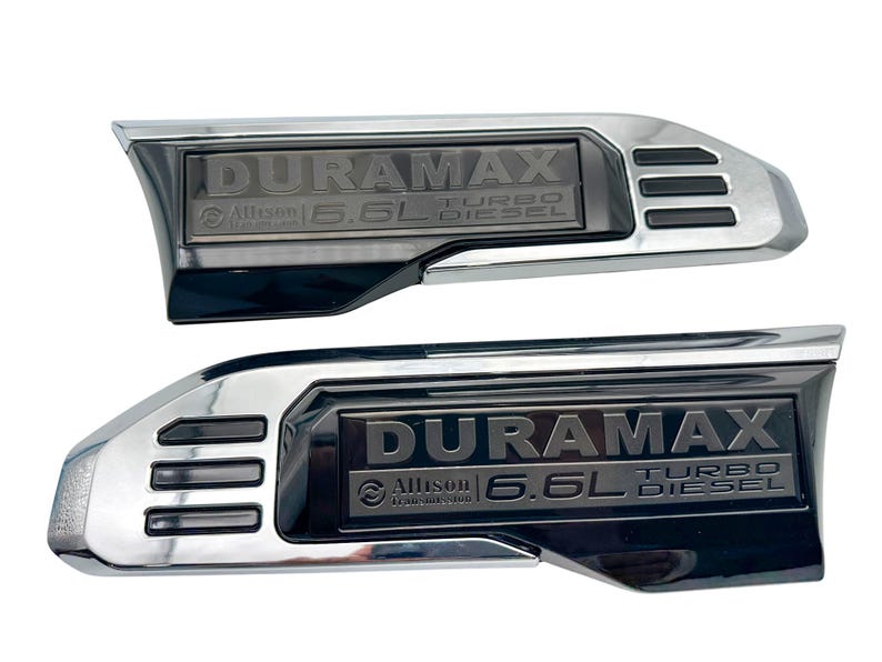 May include: Two chrome and black Duramax Turbo Diesel emblems. Each emblem features the text "DURAMAX", "Allison", and "6.6L TURBO DIESEL". The emblems have a curved shape with a rectangular centre.