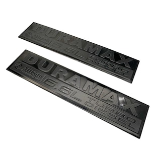 May include: Two black rectangular emblems with the text "DURAMAX 6.6L TURBO DIESEL" and "Allison Transmission" in raised lettering. The emblems are angled and appear to be made of a durable material.