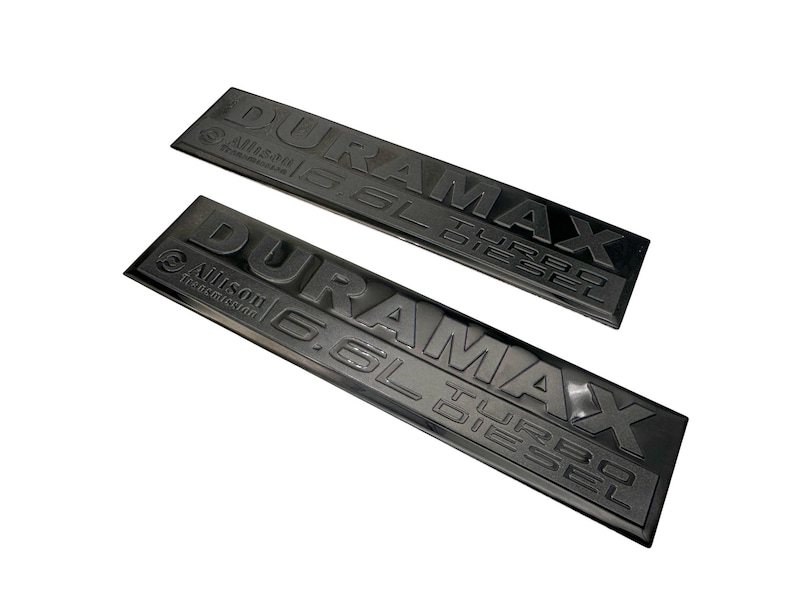 May include: Two black rectangular emblems with the text "DURAMAX 6.6L TURBO DIESEL" and "Allison Transmission" in raised lettering. The emblems are angled and appear to be made of a durable material.
