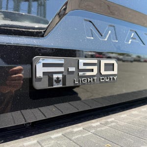 May include: Close-up of a black truck tailgate with the embossed word "MAVERICK" and an "F-150 LIGHT DUTY" emblem. The emblem includes a Canadian maple leaf. The truck's paint reflects the surrounding environment.