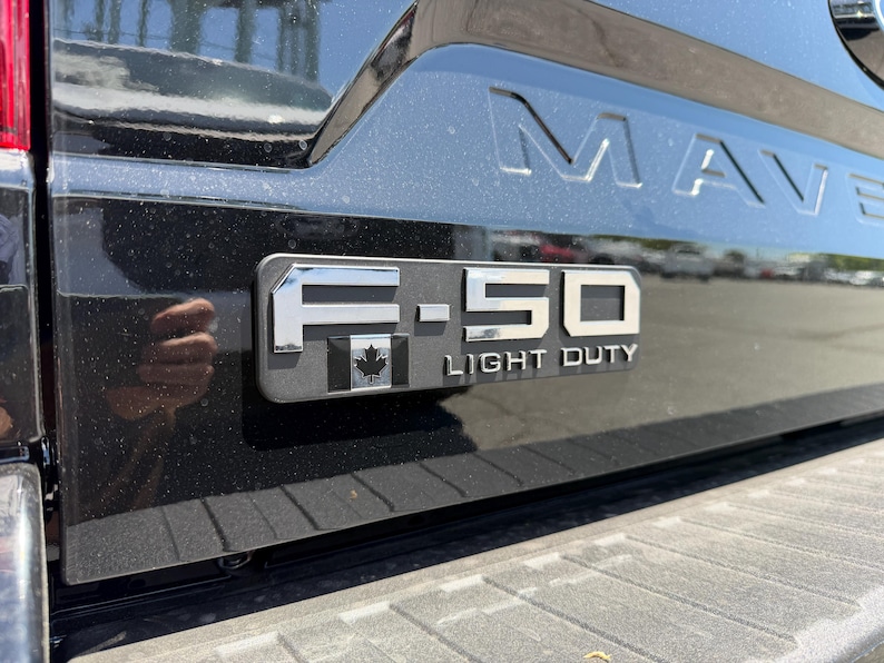 May include: Close-up of a black truck tailgate with the embossed word "MAVERICK" and an "F-150 LIGHT DUTY" emblem. The emblem includes a Canadian maple leaf. The truck's paint reflects the surrounding environment.