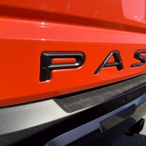 May include: Close-up of the rear of an orange vehicle with the black word "PASSPORT" across the center. The vehicle's bumper is black, and a license plate is visible in the upper right corner.