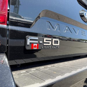 May include: Black Ford Maverick truck bed with the word "MAVERICK" embossed across the tailgate. A chrome emblem reads "F-150 LIGHT DUTY" with a Canadian flag. The Ford logo is visible.