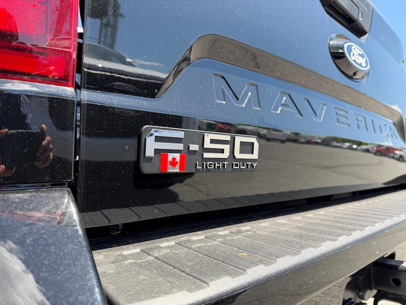 May include: Black Ford Maverick truck bed with the word "MAVERICK" embossed across the tailgate. A chrome emblem reads "F-150 LIGHT DUTY" with a Canadian flag. The Ford logo is visible.