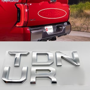 Peut inclure: Lettres chromées de l'emblème "TUNDRA". Les lettres mesurent 0,8 cm de haut et sont composées de six lettres. L'image montre les lettres sur une surface blanche et l'arrière rouge d'un camion.