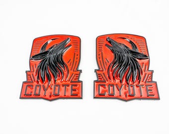May include: Two red and black metal emblems with a coyote howling at the moon. The emblems have the word "COYOTE" written in black letters on the bottom.