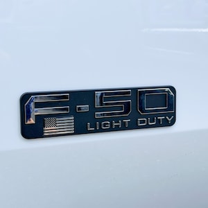 May include: A black and chrome F-150 Light Duty emblem with an American flag. The emblem is rectangular with rounded corners and is mounted on a white surface. The text "F-150" and "LIGHT DUTY" are visible.