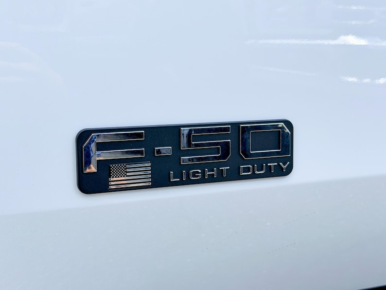 May include: A black and chrome F-150 Light Duty emblem with an American flag. The emblem is rectangular with rounded corners and is mounted on a white surface. The text "F-150" and "LIGHT DUTY" are visible.