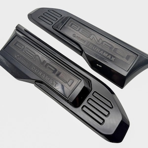 May include: Two glossy black automotive emblems featuring the text DENALI and DURAMAX in raised lettering. Each piece includes a rectangular section with three horizontal bars and a small logo emblem below the word DENALI on the surface.