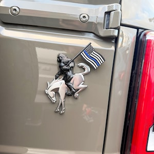 May include: A chrome emblem featuring a rider on a bucking horse, holding a flag with a blue stripe. The emblem is mounted on a tan vehicle panel, near a red taillight.