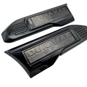 May include: Two black DURAMAX Turbo Diesel emblems. The emblems have a rectangular shape with rounded edges and feature the text "DURAMAX 6.6L Turbo Diesel" and "Allison Transmission". The emblems are glossy.