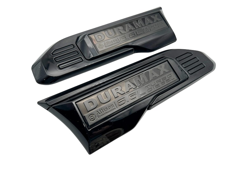 May include: Two black DURAMAX Turbo Diesel emblems. The emblems have a rectangular shape with rounded edges and feature the text "DURAMAX 6.6L Turbo Diesel" and "Allison Transmission". The emblems are glossy.