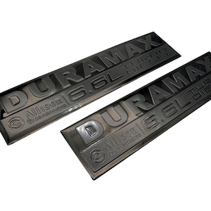 May include: Two black DURAMAX 6.6L TURBO DIESEL emblems with Allison Transmission branding. The emblems are rectangular and have raised lettering. The background is white.