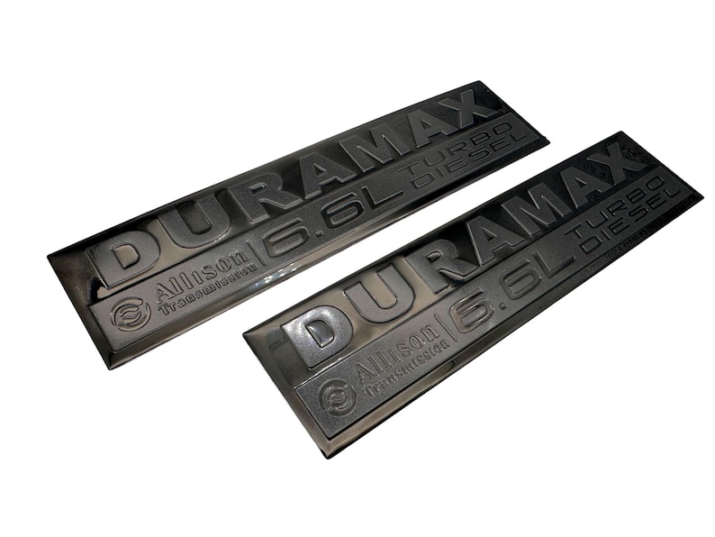 May include: Two black DURAMAX 6.6L TURBO DIESEL emblems with Allison Transmission branding. The emblems are rectangular and have raised lettering. The background is white.