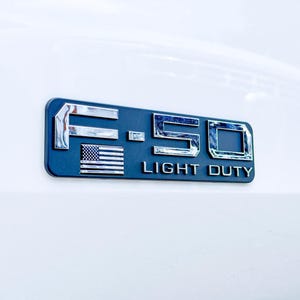 May include: A dark blue and chrome F-150 Light Duty emblem with an American flag. The emblem is rectangular and mounted on a white surface. The letters and numbers are chrome and the text "LIGHT DUTY" is in white.