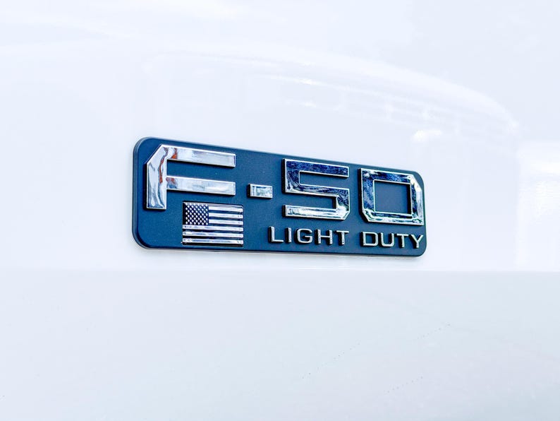 May include: A dark blue and chrome F-150 Light Duty emblem with an American flag. The emblem is rectangular and mounted on a white surface. The letters and numbers are chrome and the text "LIGHT DUTY" is in white.