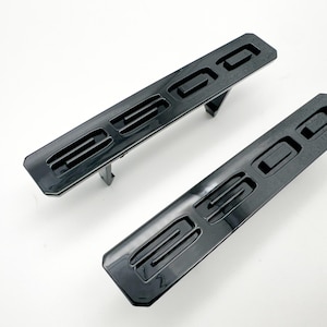 May include: Two black plastic car badges with the number "2500" embossed on them. The badges have a raised design and are intended to be attached to a vehicle.