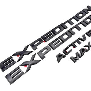 May include: A set of four black automotive emblems featuring the word EXPEDITION in a bold, sans-serif font, along with two smaller separate emblems displaying the words ACTIVE and MAX in the same matching black finish and block lettering style.