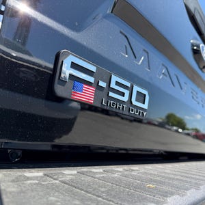 May include: Close-up of a black truck bed with the F-150 Light Duty emblem. The emblem is silver with a small American flag. The word MAVERICK is embossed on the truck bed.