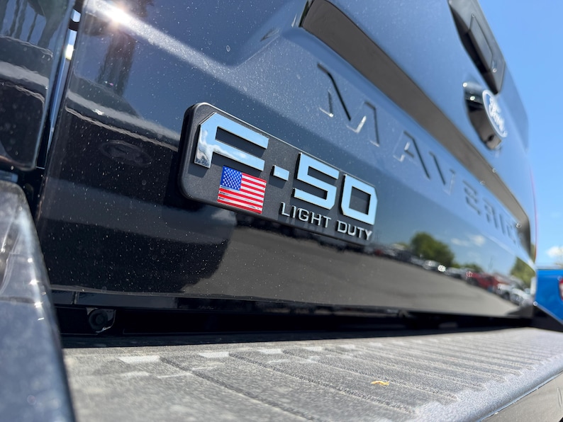 May include: Close-up of a black truck bed with the F-150 Light Duty emblem. The emblem is silver with a small American flag. The word MAVERICK is embossed on the truck bed.