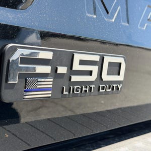 May include: A black vehicle's emblem with the text "F-150 LIGHT DUTY" in silver. The emblem includes a small American flag with a blue stripe. The background is a black, glossy surface.