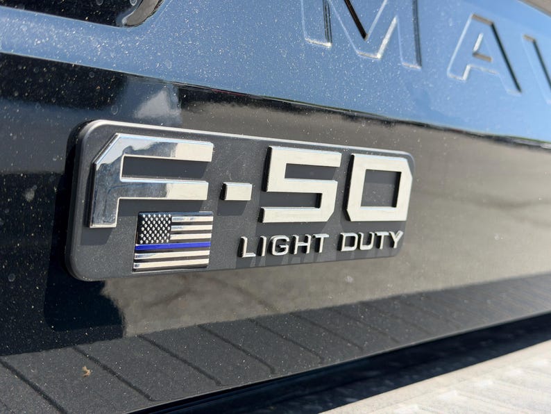 May include: A black vehicle's emblem with the text "F-150 LIGHT DUTY" in silver. The emblem includes a small American flag with a blue stripe. The background is a black, glossy surface.