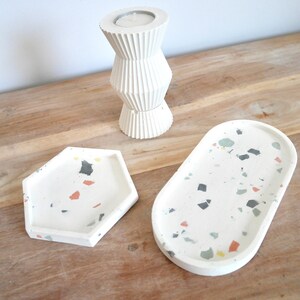 Peut inclure: Trois objets de décoration intérieure en béton blanc sur une surface en bois. Comprend un bougeoir haut et nervuré et deux plateaux terrazzo de formes hexagonales et ovales. Les plateaux ont des éclats colorés.