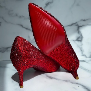 May include: A pair of red high heels covered in sparkling rhinestones. The shoes have pointed toes and stiletto heels. The soles are a solid red color. The shoes are displayed on a white and gray marble surface.