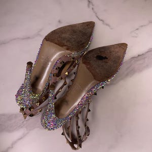 May include: A pair of high-heeled shoes with pointed toes. The shoes are adorned with iridescent rhinestones and small gold-colored studs. The heels and straps are a light brown color. The soles show signs of wear.