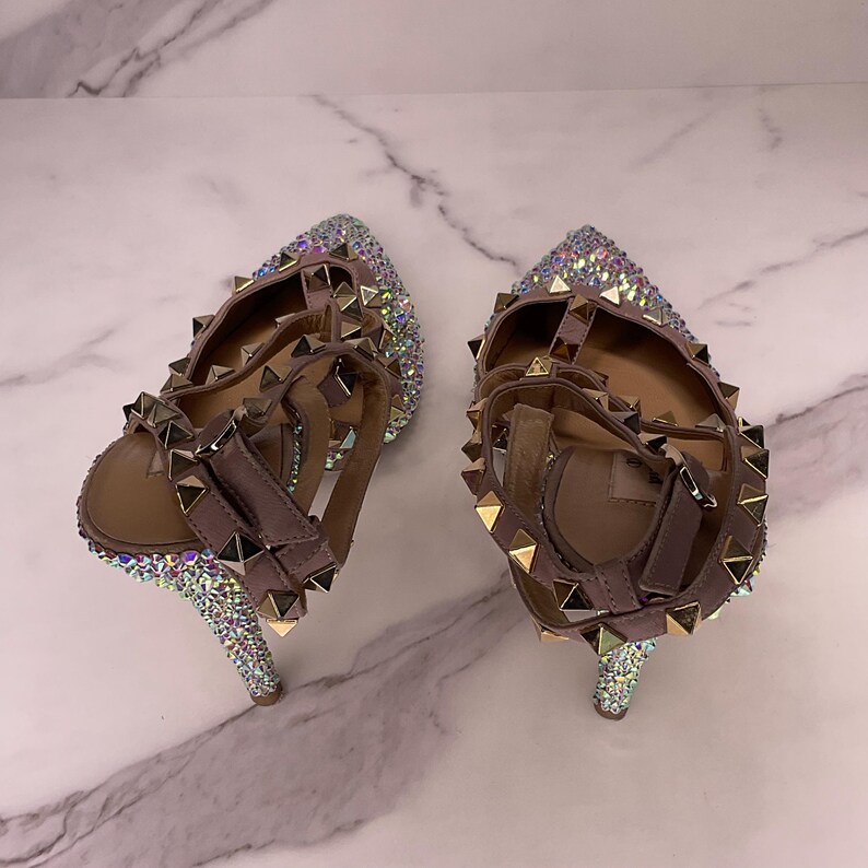 May include: A pair of high-heeled sandals with a light brown strap and a pointed toe. The sandals are embellished with iridescent rhinestones and gold-colored pyramid studs. The heels are approximately 4 inches tall.