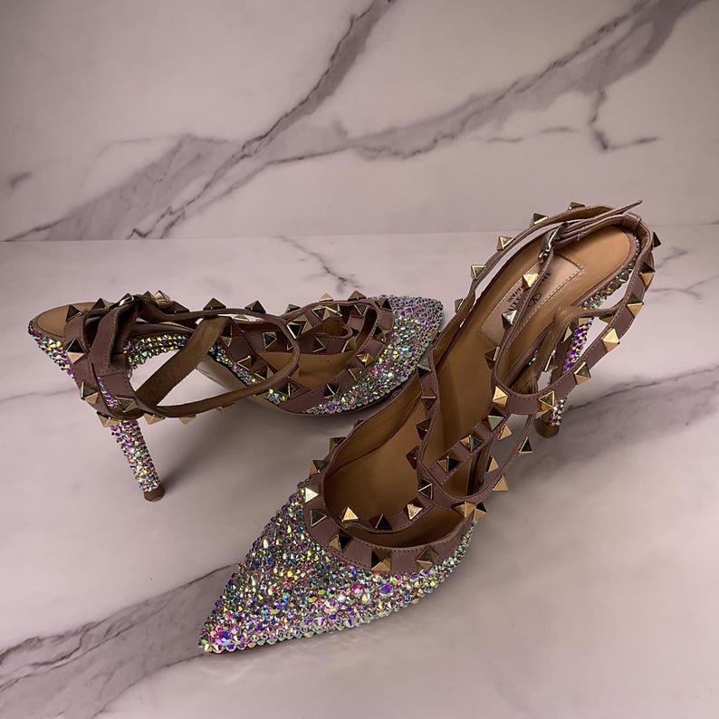 May include: A pair of mauve-colored high-heeled shoes with pointed toes. The shoes are embellished with sparkling rhinestones and gold-colored pyramid studs. The heels and straps are also adorned with studs. The shoes are set against a marble background.