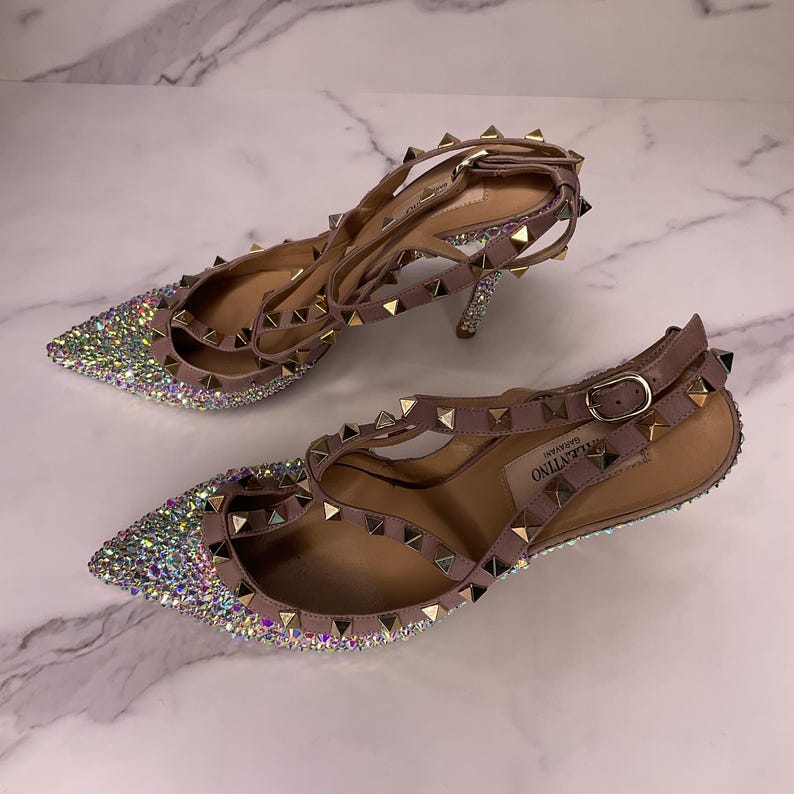 May include: A pair of high-heeled, pointed-toe shoes with a light brown strap and a silver buckle. The toe of the shoe is covered in sparkling rhinestones. The straps and heel are adorned with gold-colored studs.