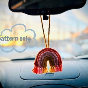 May include: A crochet rainbow car charm hanging from a rearview mirror. The rainbow is made of pink, red, orange, yellow, and green yarn. The charm has tassels in pink, yellow, and orange. The text "pattern only" is written in a thought bubble.